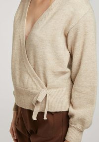 Cardigan wrap beige en matériau doux et texturé avec des poignets à côtes et une taille nouée. Présente un décolleté en V profond et une coupe confortable.