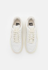 White leather sneakers with perforated toe area, light gray accents, and cream-colored rubber soles. Laces are beige with a logo patch on the tongue.