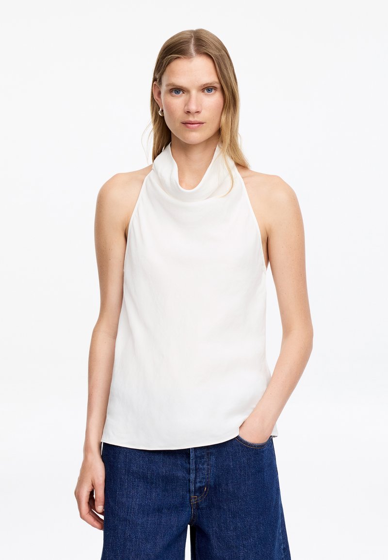 Femme aux cheveux blonds raides portant un haut blanc sans manches à col bénitier et un jean large bleu foncé, une main dans la poche.