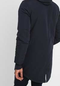 Person som bär en lång, mörk marinblå huva kappa sedd bakifrån, med en liten slits i nederkanten.