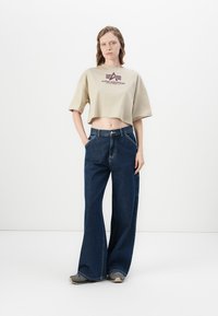 Beige cropped t-skjorte med "ALPHA INDUSTRIES" logo, kombinert med mørkeblå vide jeans. Modellen står med hendene på hoftene, mot en nøytral bakgrunn.
