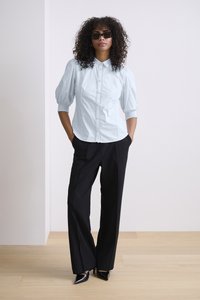 Blusa azzurro chiaro con maniche a sbuffo e dettagli in bottoni, abbinata a pantaloni neri a gamba larga e décolleté neri a punta, su uno sfondo neutro.