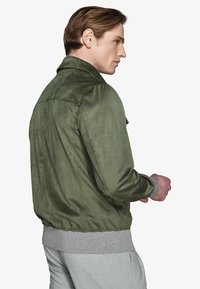 Homme tourné de côté portant une veste en suède verte par-dessus un pull gris et un pantalon gris clair sur un fond blanc.