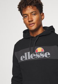 Schwarze Kapuzenjacke mit einem grauen Streifen und dem "ellesse"-Logo sowie einem bunten Emblem. Enthält Kordeln mit strukturierten Griffen.