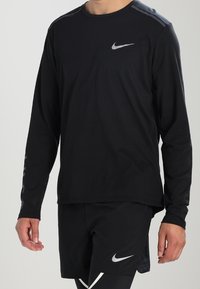 Schwarzes Langarm-Nike-Sporthemd mit Rundhalsausschnitt, das ein silbernes Logo auf der Brust und Details an den Schultern aufweist. Der Stoff wirkt glatt und leicht.