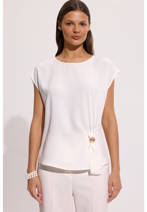 Kobieta nosząca białą, bezrękawnikową bluzkę z ozdobnym złotym guzikiem po boku oraz dopasowane, białe spodnie.