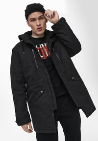 Svart parka jacka med huva, med dragkedjefickor, justerbara snoddar och avslappnad passform. Bärs över en svart grafisk tröja.