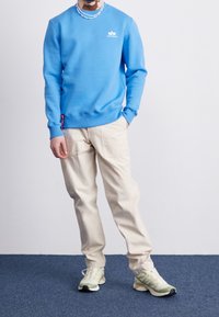 Persona vestida con sudadera azul, pantalones beiges y zapatillas ligeras verdes y blancas, de pie sobre una alfombra azul contra un fondo blanco.