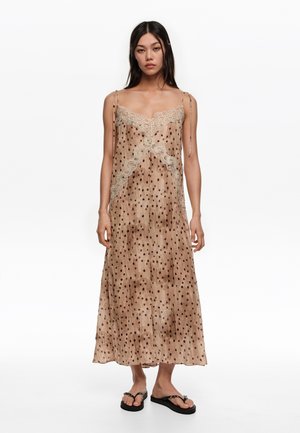 Kobieta ubrana w beżową, bezrękawnikową sukienkę midi z cienkimi ramiączkami, w czarne kropki, z koronkowymi wykończeniami, w czarnych platformowych japonkach, stojąca na białym tle.