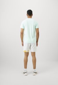 Mann steht mit dem Rücken zur Kamera, trägt ein hellblaues Hemd, weiße Shorts mit gelbem Muster, weiße Socken und weiße Sneakers vor einem schlichten Hintergrund.
