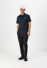 Jūrine polo marškinėliai su trumpomis rankovėmis ir geltonu logotipu; derinami su tamsiais krovinių džinsais ir juodais batais; modelis dėvi šviesios spalvos kepurę.