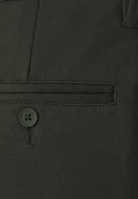 Ciemnozielony materiał z guzikem poniżej poziomej kieszeni ze szwem, pokazujący szczegółowe przeszycia na elemencie odzieży.