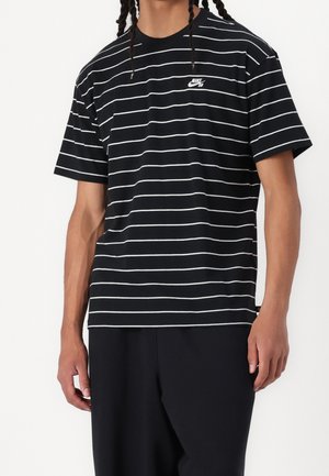 Osoba nosząca czarną koszulkę z krótkim rękawem z białymi poziomymi paskami i małym białym logo Nike SB na lewej piersi.
