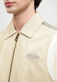 Beige Lederjacke mit grünem Akzent, Reißverschluss, Spitzkragen und gesticktem "GUESS"-Logo. Glatte Textur, modernes Design.