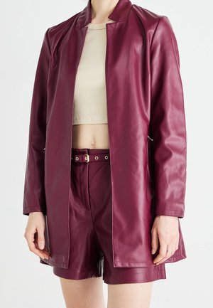 Femme portant une veste en faux cuir bordeaux et un short assorti avec ceinture, ainsi qu'un crop top beige, debout devant un fond blanc uni.