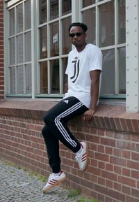 Balta Juventus logo T-kreklu, melnas Adidas sporta biksēs ar baltām svītrām, sarkani un balti sporta apavi. Pilsētas vide ar ķieģeļu sienu un logiem.