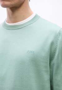 Gros plan sur une personne portant un pull vert clair à col rond avec le mot « BOSS » brodé sur la poitrine, par-dessus un sous-pull blanc.