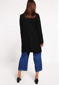 Svart, lång kappa med en jämn textur, skräddarsydd passform och mjukt tyg. Kombineras med vida blå denimbyxor och svarta klacksandaler.