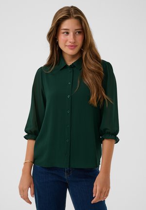 Frau mit langem welligem Haar, die eine dunkelgrüne Hemdbluse mit Dreiviertelärmeln und blaue Jeans vor einem einfarbigen Hintergrund trägt.