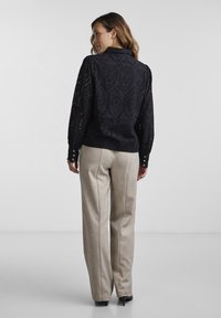 Black eyelet blouse with a textured pattern, long sleeves, and buttoned cuffs, paired with light beige tailored trousers and high heels.