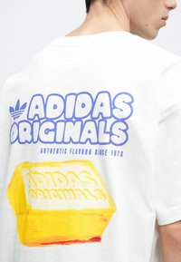 Balta kokvilnas T-krekls ar izteiksmīgu zilu "ADIDAS ORIGINALS" grafiku un dzeltenu sviesta bloka ilustrāciju aizmugurē. Mīksta tekstūra.