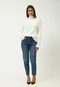 White knitted turtleneck sweater with a cable pattern, paired with blue fitted jeans and black high-heeled shoes. Plain background.