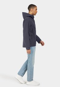 Marinblå vattentät jacka med huva, framficka med dragkedja och kardborreknäppningar, kombinerad med ljusblå jeans och vita sportskor.