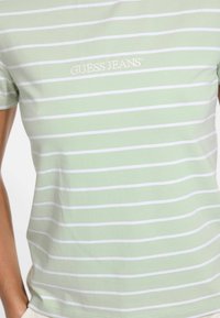 Zaļš svītrains t-krekls no maiga auduma, ar baltām horizontālām svītrām un izšūtu "GUESS JEANS" logo baltā krāsā. Īsas piedurknes, pieguļošs griezums.