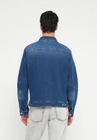 Mann trägt eine blaue Jeansjacke und hellgraue Jeans, steht mit dem Rücken zur Kamera vor einem einfachen weißen Hintergrund.