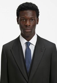 Abito nero con finitura testurizzata, camicia bianca e cravatta blu a motivo. Dettaglio accentuato rosso visibile sulla rever.