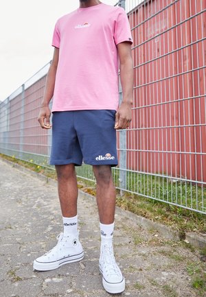 Persoon draagt een roze ellesse t-shirt, navy ellesse shorts, witte ellesse sokken en witte hoge sneakers, staand bij een metalen hek buiten.