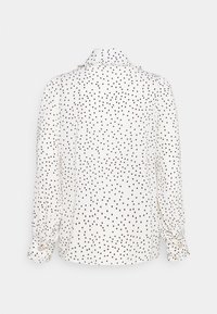 Blouse blanche à manches longues avec des pois noirs, poignets boutonnés et un col haut à volants, vue de dos sur un fond uni.
