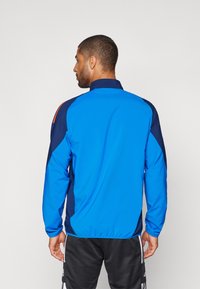Chaqueta azul y marino con cuello alto, mangas largas y corte atlético. Textura suave con paneles laterales en contraste y acentos naranjas.