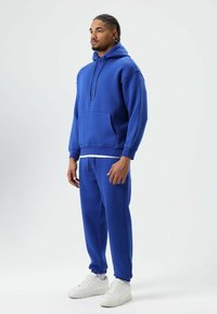 Blue hoodie with a front pocket and drawstring hood, paired with matching blue joggers, set against a light background. White sneakers.