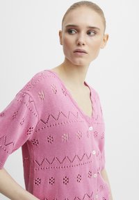 Ružový pletený kardigán s krátkymi rukávmi, s výstrihom do V, zapínaním na gombíky a dekoračnými otvorenými vzormi po celom povrchu.