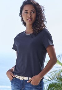 Mornarsko modra majica s kratkimi rokavi in okroglim izrezom, okrašena s srebrnimi dekorativnimi žebljički na ovratniku, nosi se z pletenim belim pasom.