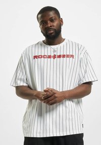 T-shirt blanc à manches courtes avec des rayures verticales noires, arborant un logo rouge "ROCA WEAR" sur la poitrine. Matière en coton, coupe décontractée.