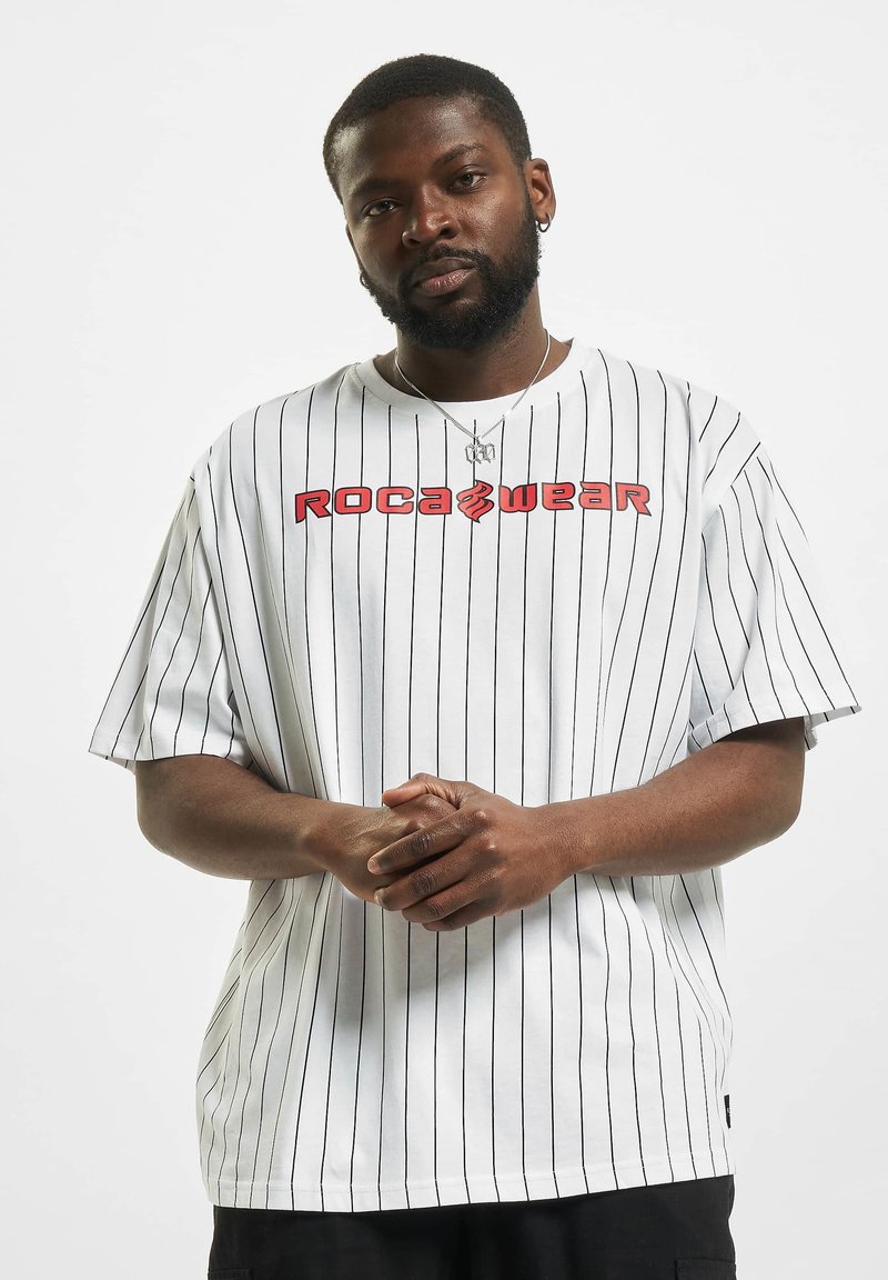 T-shirt blanc à manches courtes avec des rayures verticales noires, arborant un logo rouge "ROCA WEAR" sur la poitrine. Matière en coton, coupe décontractée.