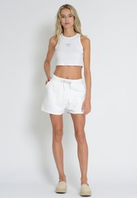 White cropped tank top with light branding, paired with white drawstring shorts featuring side pockets and a soft texture. Light-colored footwear.