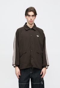 Καφέ μπουφάν με κουμπιά μπροστά, διακοσμημένο με λευκές τρεις ρίγες στα μανίκια και κεντημένο λογότυπο στο στήθος.