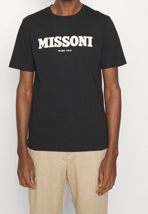 Persoon draagt een zwart Missoni T-shirt met witte tekst "MISSONI MILANO - ITALIA" en beige broek, staand tegen een effen witte achtergrond.