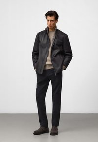 Mężczyzna stoi z rękami w kieszeniach, ubrany w czarną skórzaną kurtkę, beżowy sweter, czarne spodnie i ciemnobrązowe buty na tle jednolitego tła.