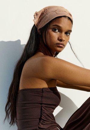 Donna con lunghi capelli neri che indossa un top marrone senza spalline e un foulard rosa ricamato, seduta davanti a uno sfondo bianco.