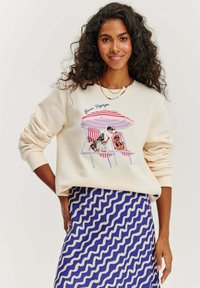 Krémová mikina se grafikou psa a textem "Bon Voyage", doplněná modrou sukni s vlnkovaným vzorem, představující ležérní vrstvený vzhled.