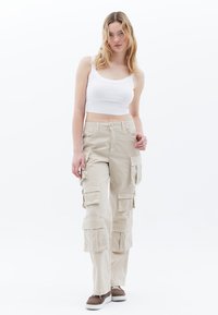 Beige cargo kalhoty s několika postranními kapsami, volný střih a lehký materiál. V kombinaci s bílým cropped topem a hnědými botami.