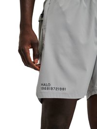 Ciemnoskóra ręka spoczywająca w pobliżu bocznej kieszeni jasnoszarych krótkich spodenek z nadrukiem "HALO 196819721981" blisko krawędzi.