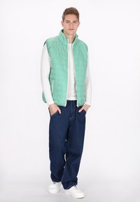 Hellgrüne Steppweste mit Stehkragen, Reißverschluss und zwei seitlichen Taschen, getragen über einem weißen Langarmshirt und dunkelblauen Jeans.