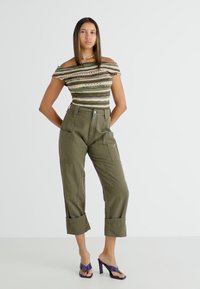 Juostuotas off-the-shoulder topas žemės atspalvių, derinamas su alyvuogių žalios spalvos plačiomis krovinių kelnėmis, turinčiomis dideles kišenes ir sukniubtais rankovėmis. Purpurinės aukštakulnės sandalai.
