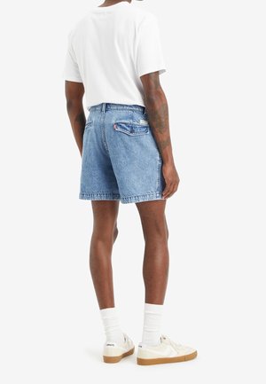 Shorts en jean bleu à coupe décontractée, ourlet brut et deux poches arrière. Portés avec un t-shirt blanc et des baskets de couleur claire.