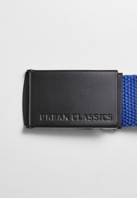 Mėlynas nertas diržas su stačiakampiu juodu metaliniu užsegimu, ant kurio išgraviruotas tekstas "URBAN CLASSICS". Tvirtas, paprastas dizainas, tinkantis laisvalaikio aprangai.